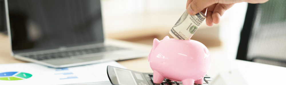 laptop on desk and hand putting money into the piggy bank sitting on top of a calculator