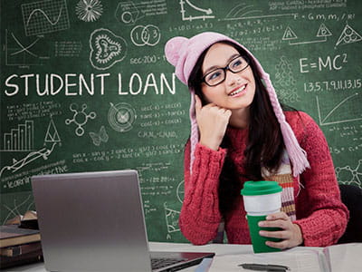Female student smiling while sitting at a desk in front of a chalkboard in her college classroom that says Student Loan