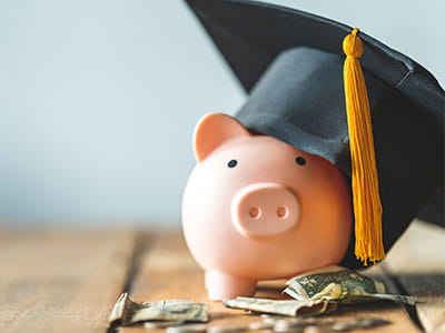 Piggy bank wearing graduation cap with cash and coins in front to convey concept of paying for college